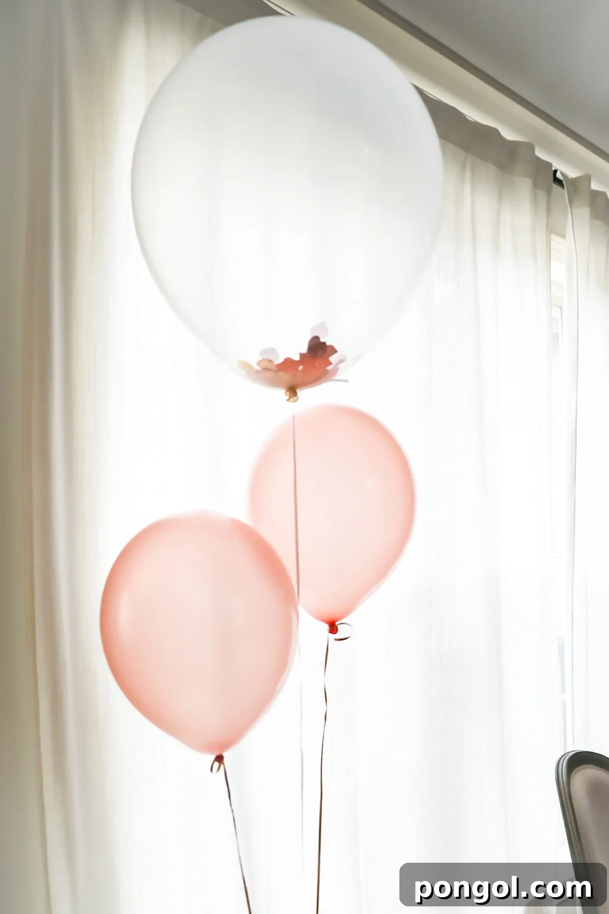 Three balloons - two pink, one clear and filled with confetti - hovering in front of a window.