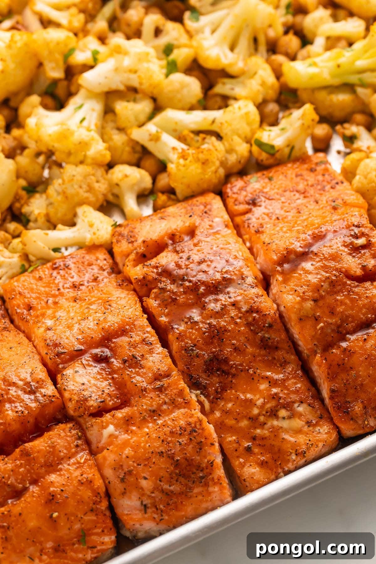 Close-up shot of four beautifully glazed hot honey salmon fillets resting on a bed of roasted cauliflower florets and chickpeas on a sheet pan, highlighting the texture and rich color of the dish.
