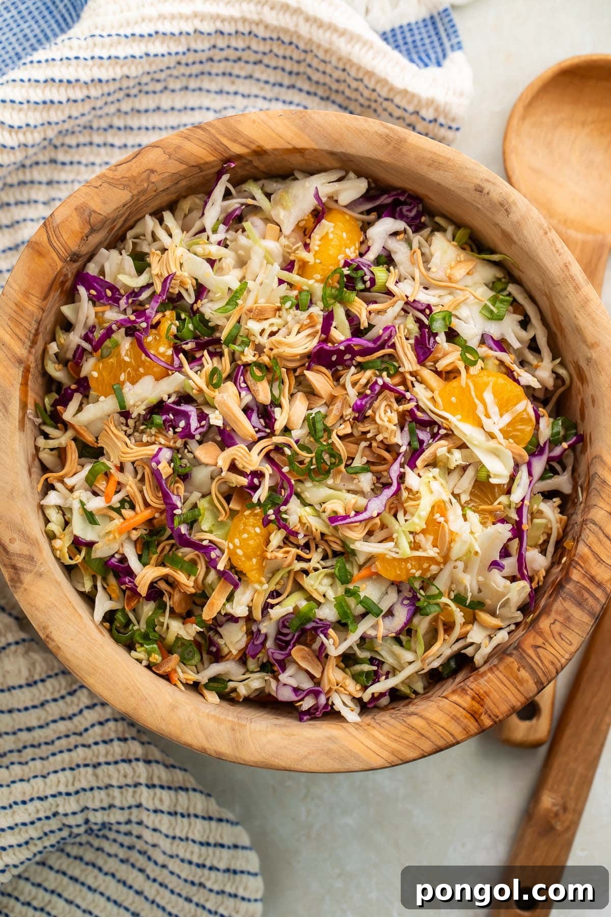 Sesame Ginger Slaw 2 A vibrant wooden bowl of Asian slaw, brimming with colorful vegetables, roasted ramen noodles, and mandarin orange segments, rests on a rustic wooden table beside a wooden spoon and a blue and white striped kitchen towel, ready to be served.