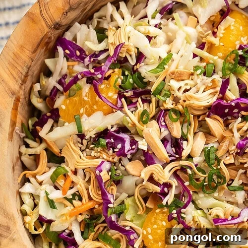 Close-up of a wooden bowl of Asian slaw, with a focus on the fresh ingredients and crunchy toppings, on a table next to a wooden spoon and a blue and white striped kitchen towel.