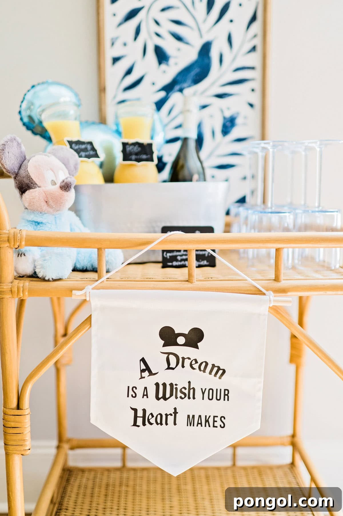 A beverage cart themed for a Magic Kingdom birthday party, with a serve-yourself mimosa bar, decorated with a pastel baby Mickey Mouse plush and a banner that reads "A dream is a wish your heart makes".