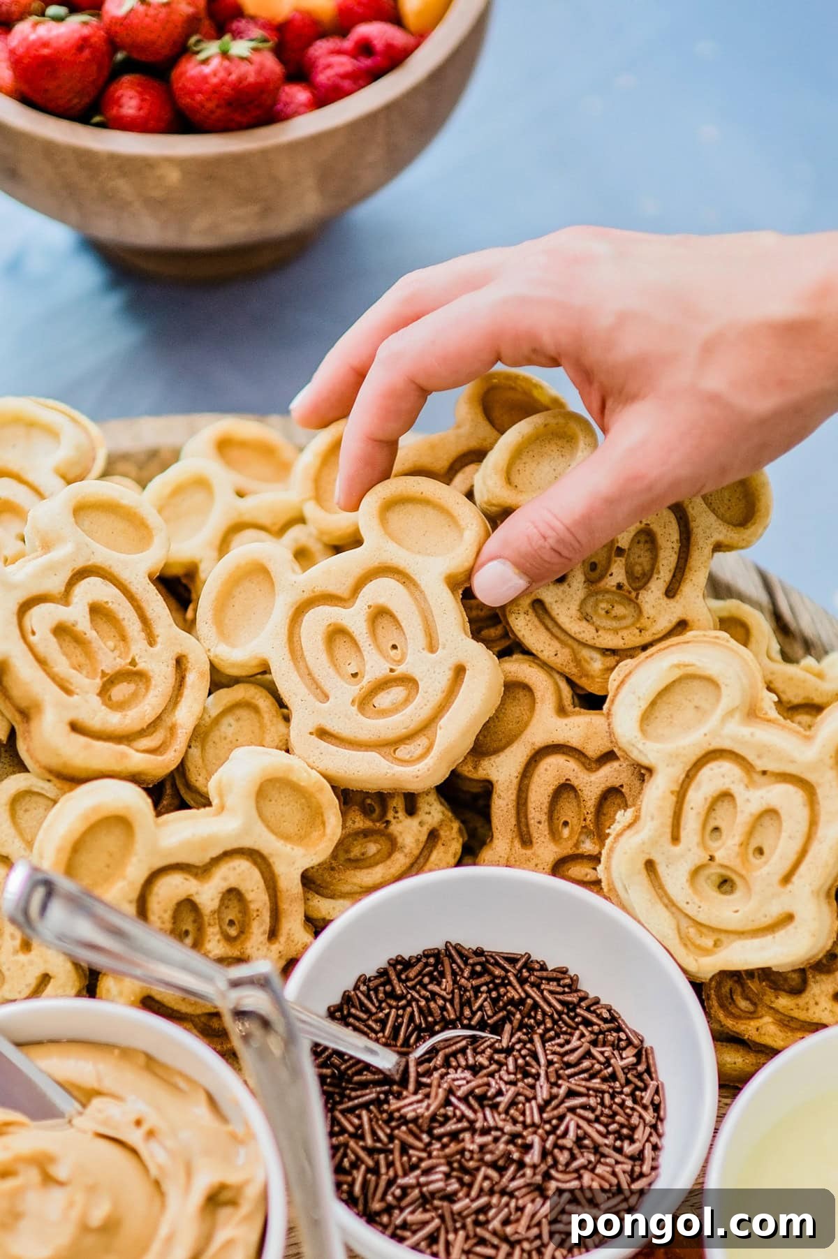 A white woman's hand reaches for a Mickey Mouse shaped waffle on a large tray with waffle toppings.