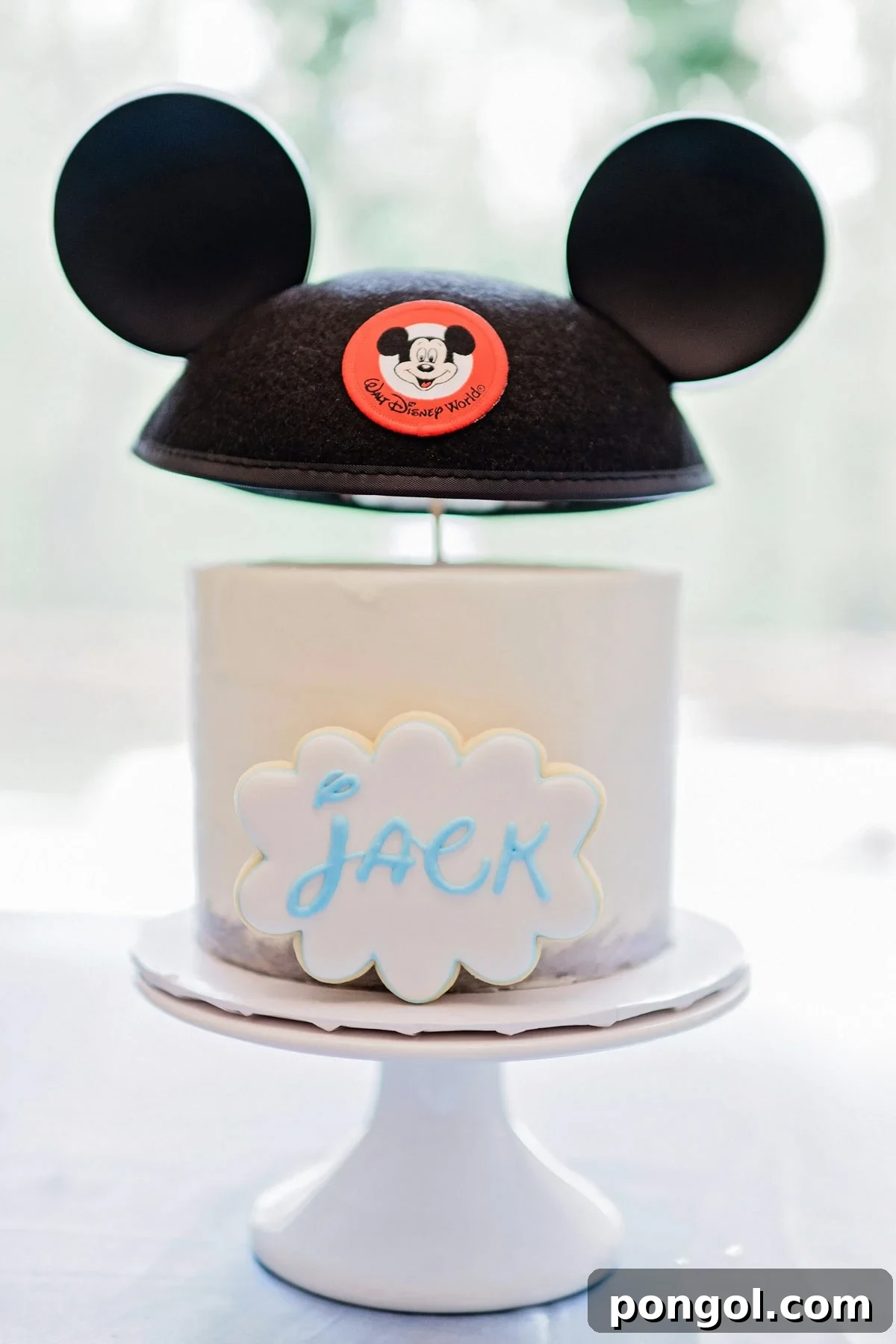 A white round birthday cake topped with a Disneyland classic ear hat. A cookie that says "Jack" in the classic Disney font leans against the front of the cake.