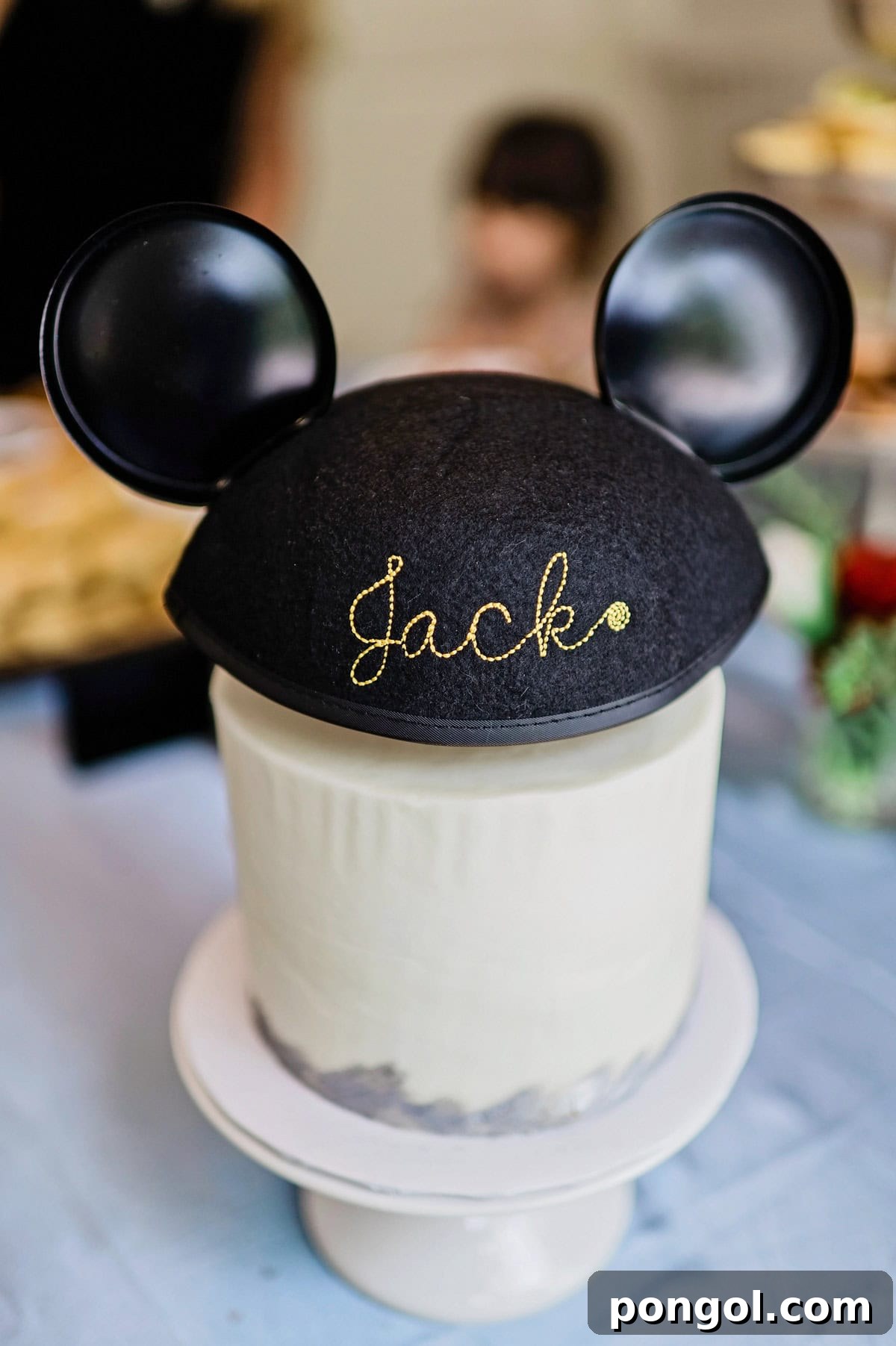 A white round birthday cake topped with a Disneyland classic ear hat. The back of the hat shows cursive gold embroidery spelling out the name "Jack".