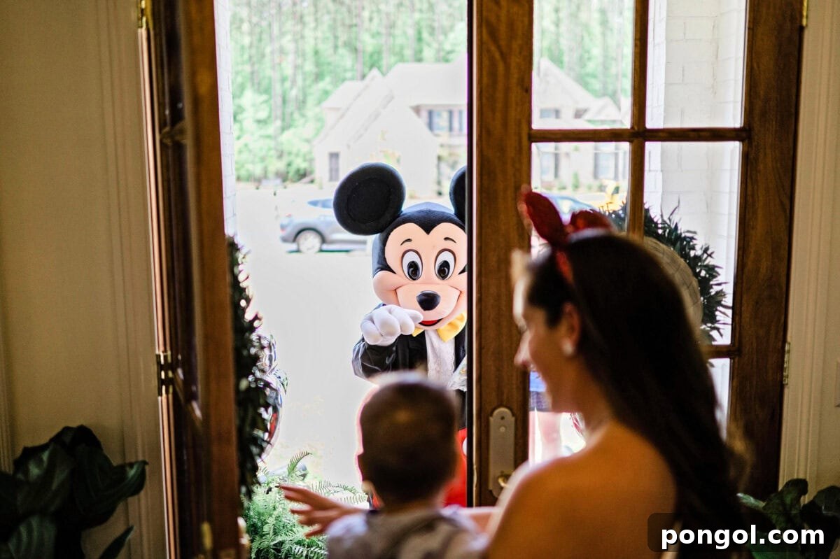 A Mickey Mouse birthday party character arrives at the front door of a house and is greeted by a woman wearing mouse ears and holding a small child.