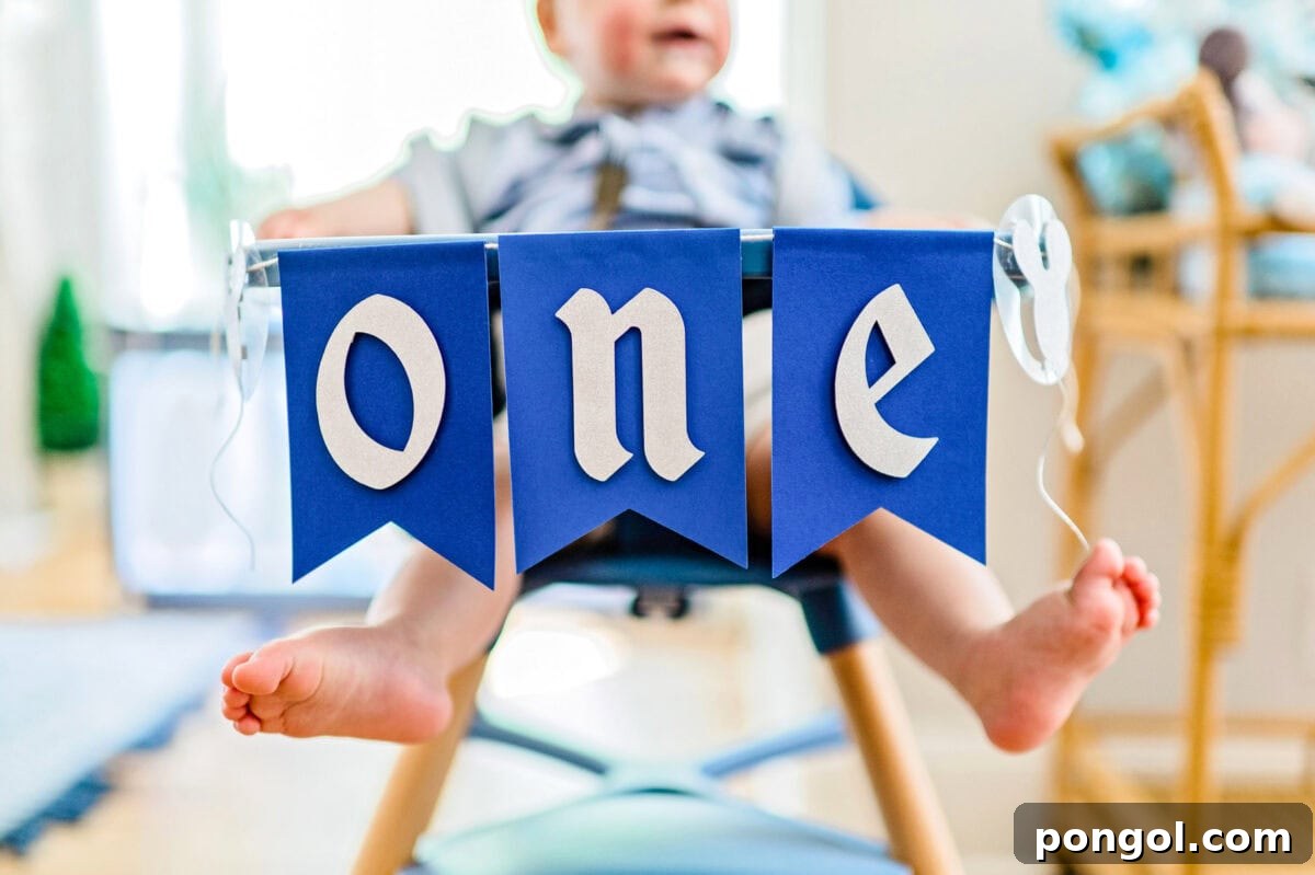 A "ONE" birthday banner hangs on the front of a high chair that a little boy is sitting in.