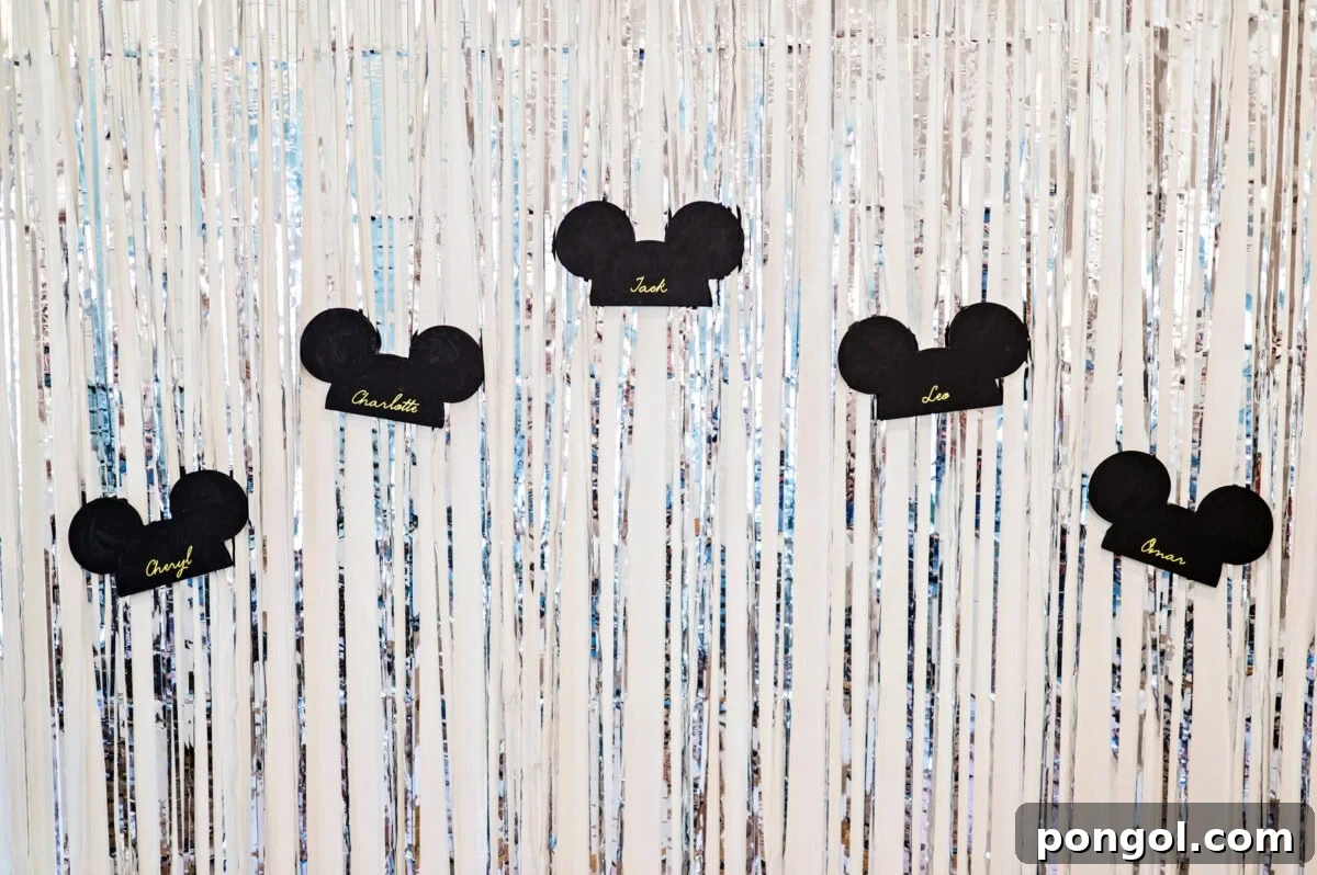 Silver streamers hang on a beige wall. 5 paper Disneyland mouse ear hats are stuck to the streamers. Each hat features the name of a person.