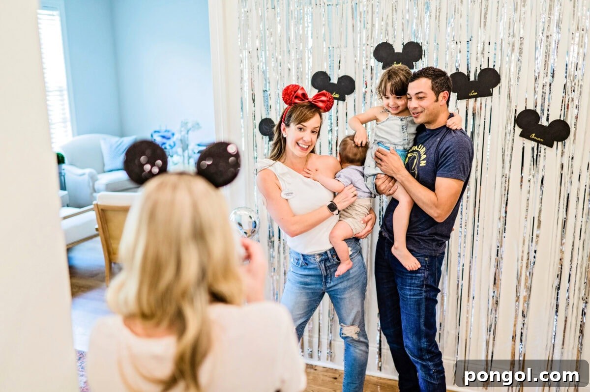 A man and a woman holding two small children pose for a photo in front of silver streamers hanging on a wall.