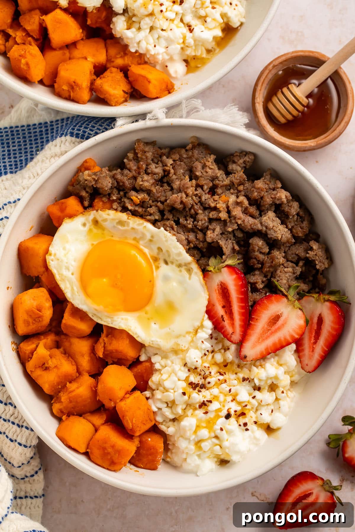 A vibrant breakfast bowl featuring creamy cottage cheese, perfectly roasted sweet potatoes, savory breakfast sausage crumbles, and fresh strawberry slices, all topped with a sunny-side-up fried egg.