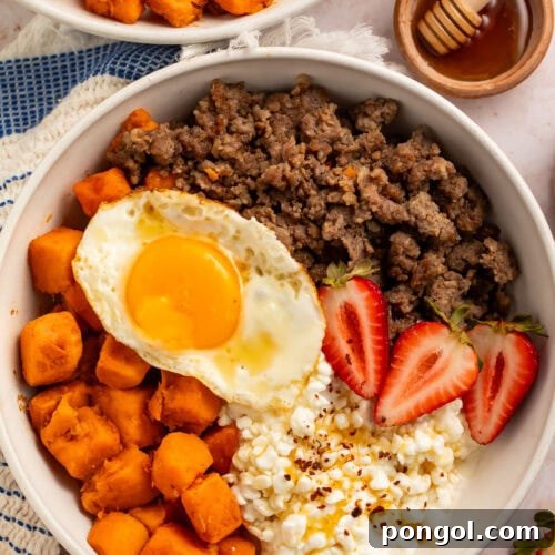 A breakfast bowl with cottage cheese, sweet potatoes, breakfast sausage, and sliced strawberries, topped with a fried egg.