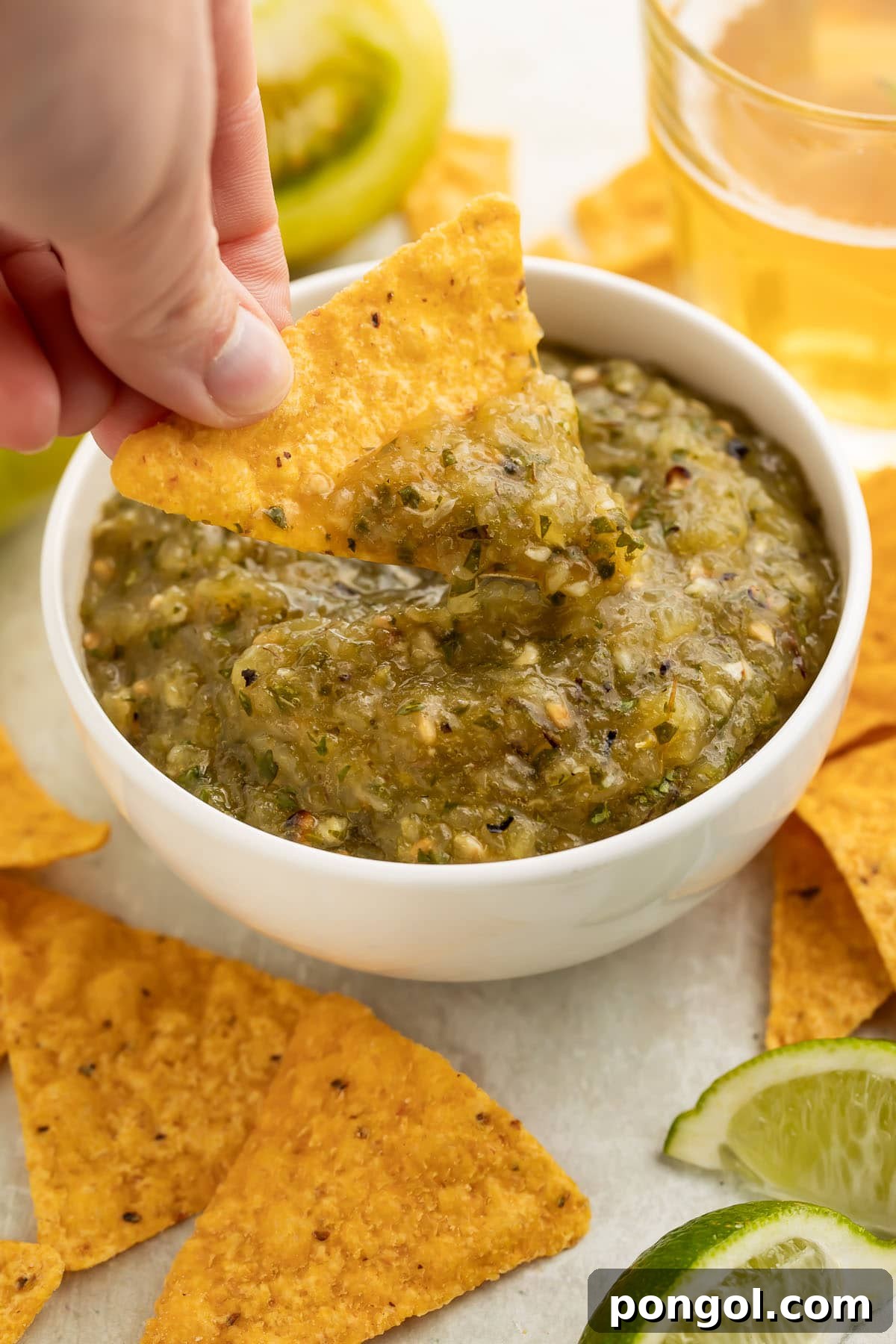 A woman's hand expertly scoops vibrant green tomato salsa with a white corn tortilla chip from a rustic white bowl.