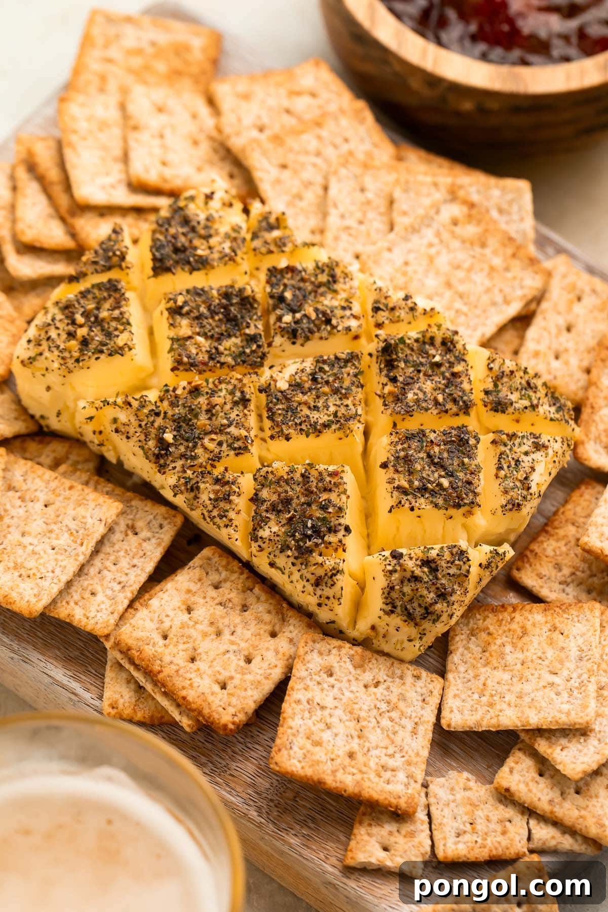 A block of smoked cream cheese, beautifully seasoned and scored with a criss-cross pattern, surrounded by crispy square wheat crackers on a rustic wooden board.