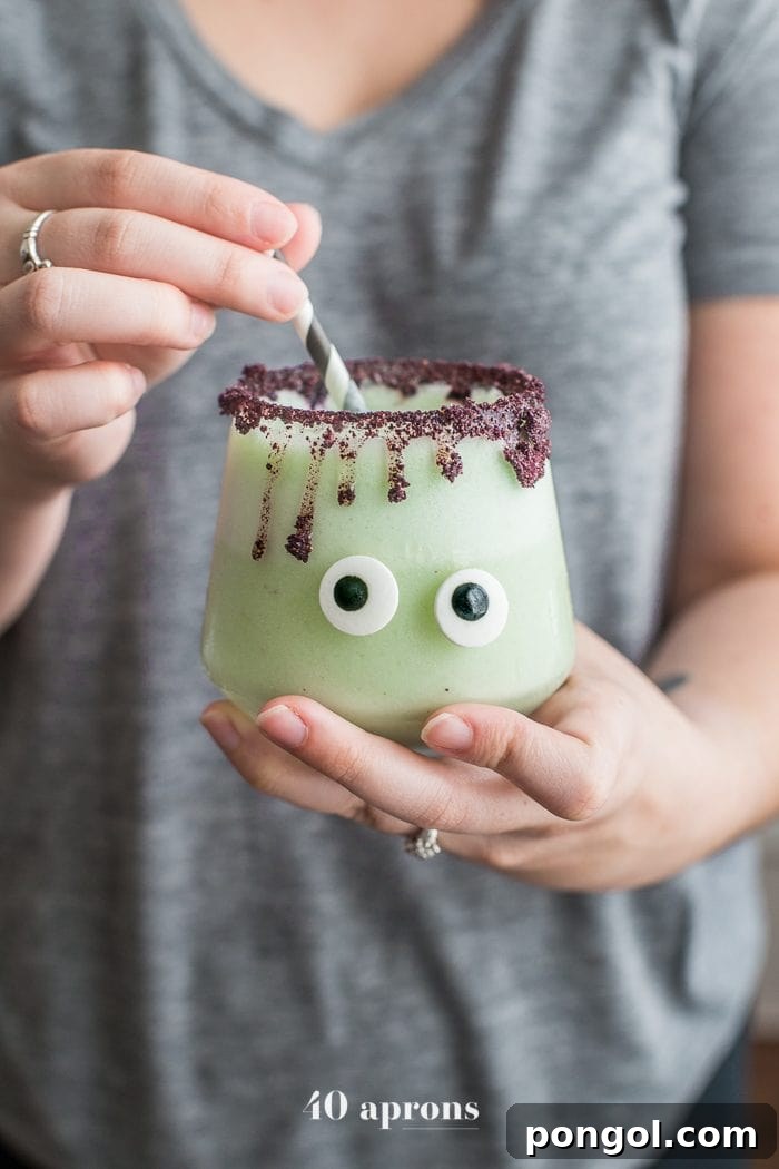 Someone holding Monsteritas green Halloween cocktails with purple dripping rim and candy monster eyes in glasses with a black and white straw