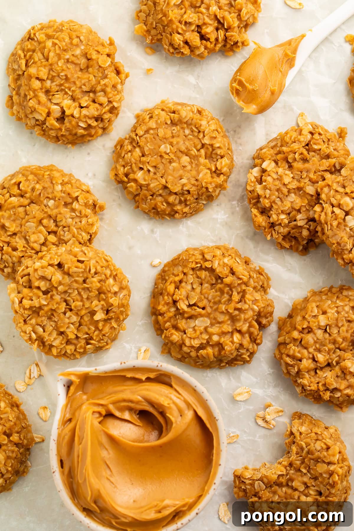 Peanut butter oat cookies on a piece of parchment paper with a measuring cup of smooth peanut butter and a white spoonful of peanut butter.