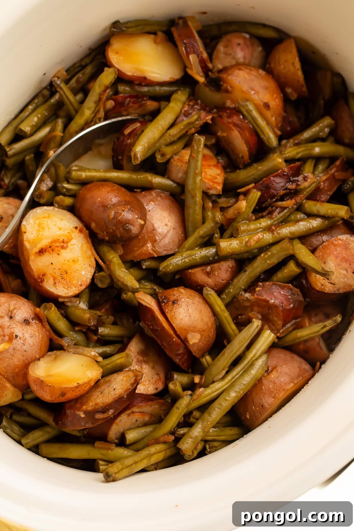 White slow cooker filled with cooked swamp potatoes showing tender red potatoes, green beans, and sliced sausage with serving spoon