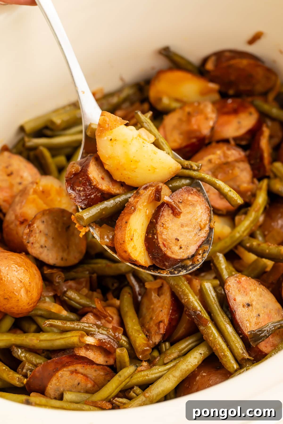 Close-up of serving spoon holding swamp potatoes with golden potato pieces, green beans, and sausage slices