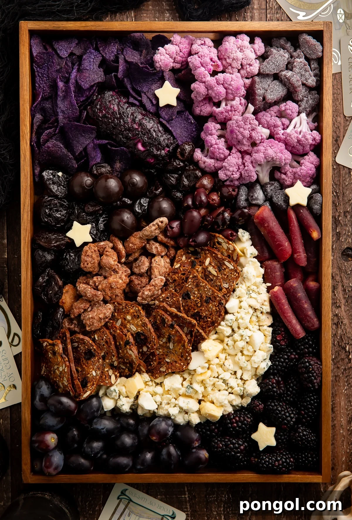 Mystic Lunar Charcuterie Board 2 Dramatic Halloween themed charcuterie board in a rustic wooden tray, featuring a crescent moon made of white cheeses, surrounded by dark purple and black fruits, vegetables, and snacks, with scattered white star cheese cutouts.