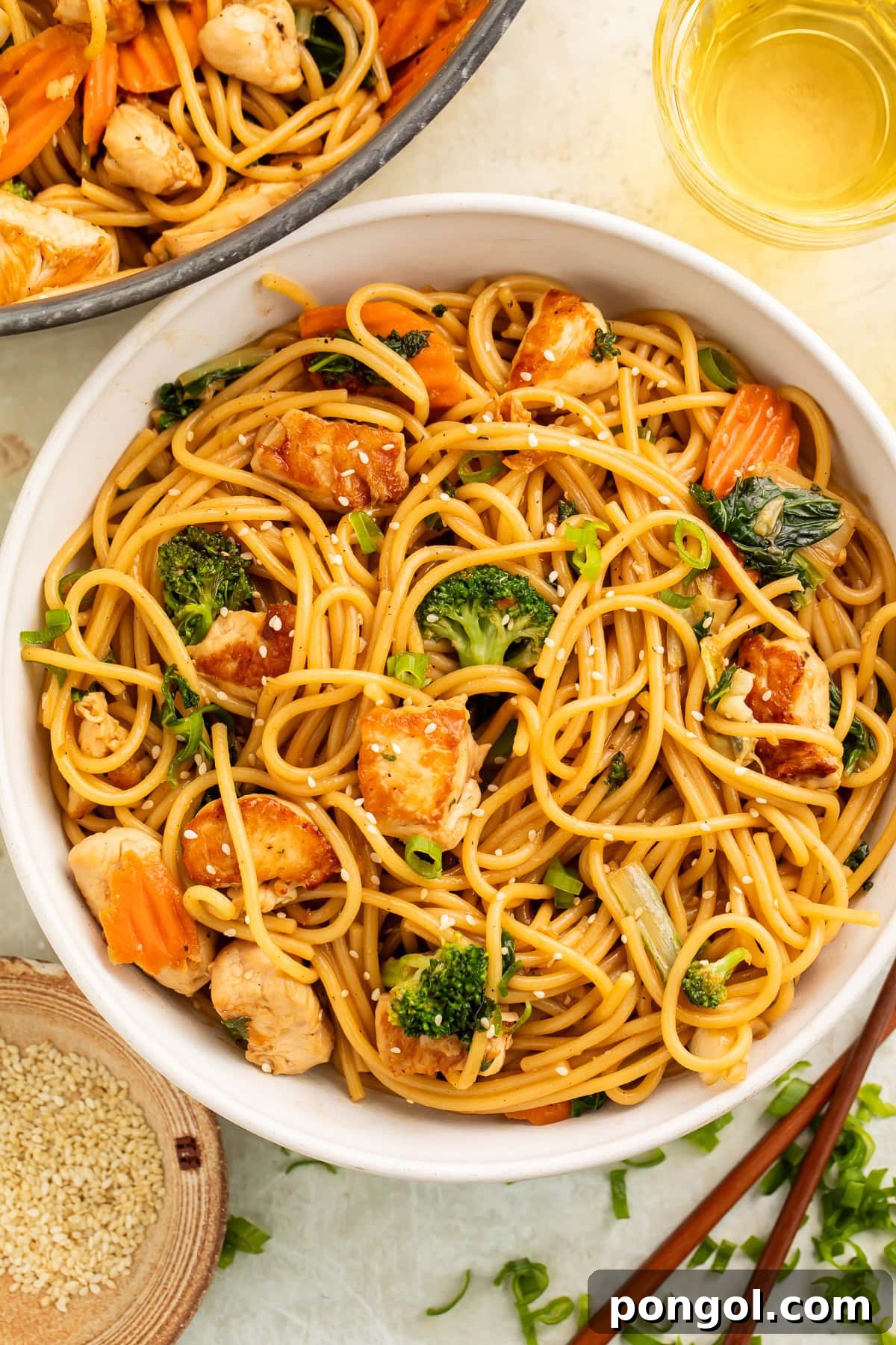 A beautifully presented bowl of sesame garlic chicken lo mein, featuring tender chicken, crisp vegetables, and glossy noodles, garnished with a sprinkle of sesame seeds and fresh green onions. Chopsticks rest alongside the bowl, inviting you to enjoy this delicious meal.