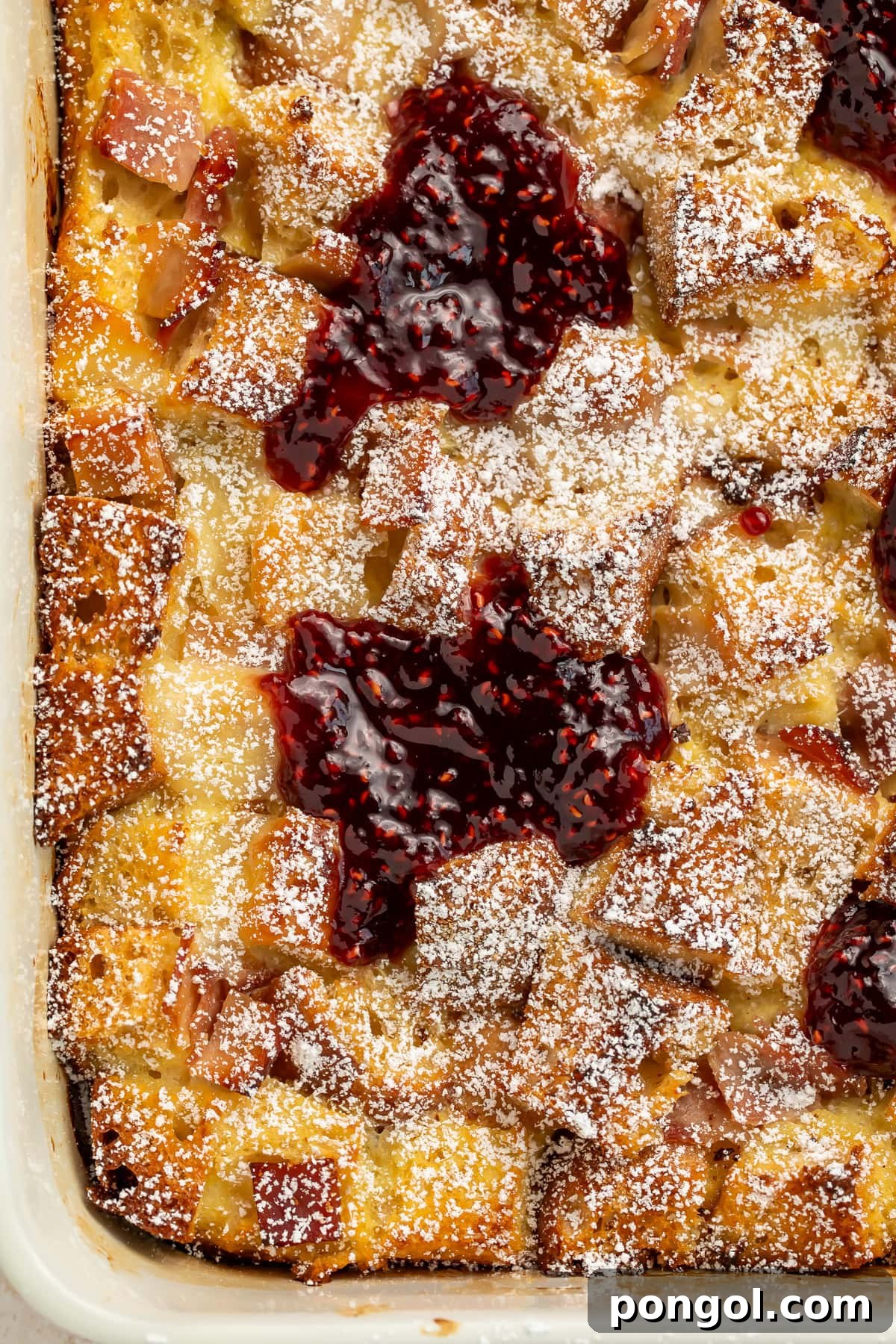 Golden-brown Monte Cristo breakfast casserole topped with powdered sugar and raspberry preserves in a white baking dish.