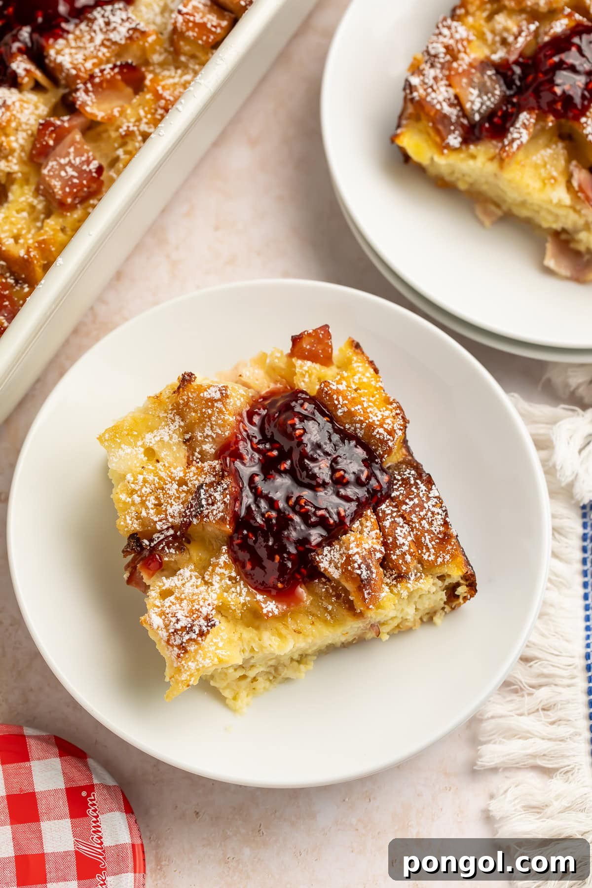 Slice of Monte Cristo breakfast casserole on a white plate topped with raspberry jam and powdered sugar.
