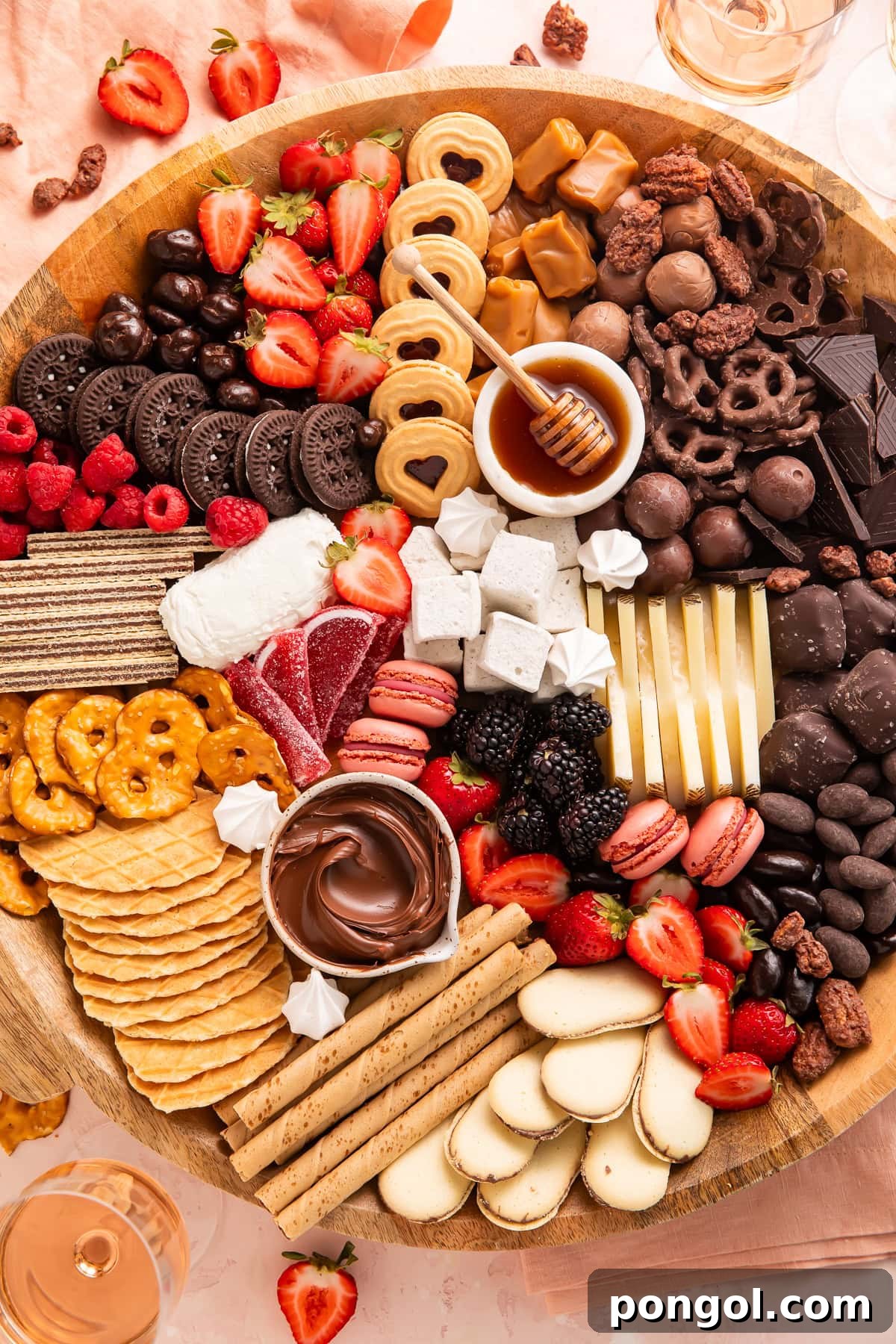 Overhead view of dessert charcuterie board with chocolates, cookies, fresh strawberries, raspberries, blackberries, cheese, honey, pretzels, and waffle cookies on wooden board.