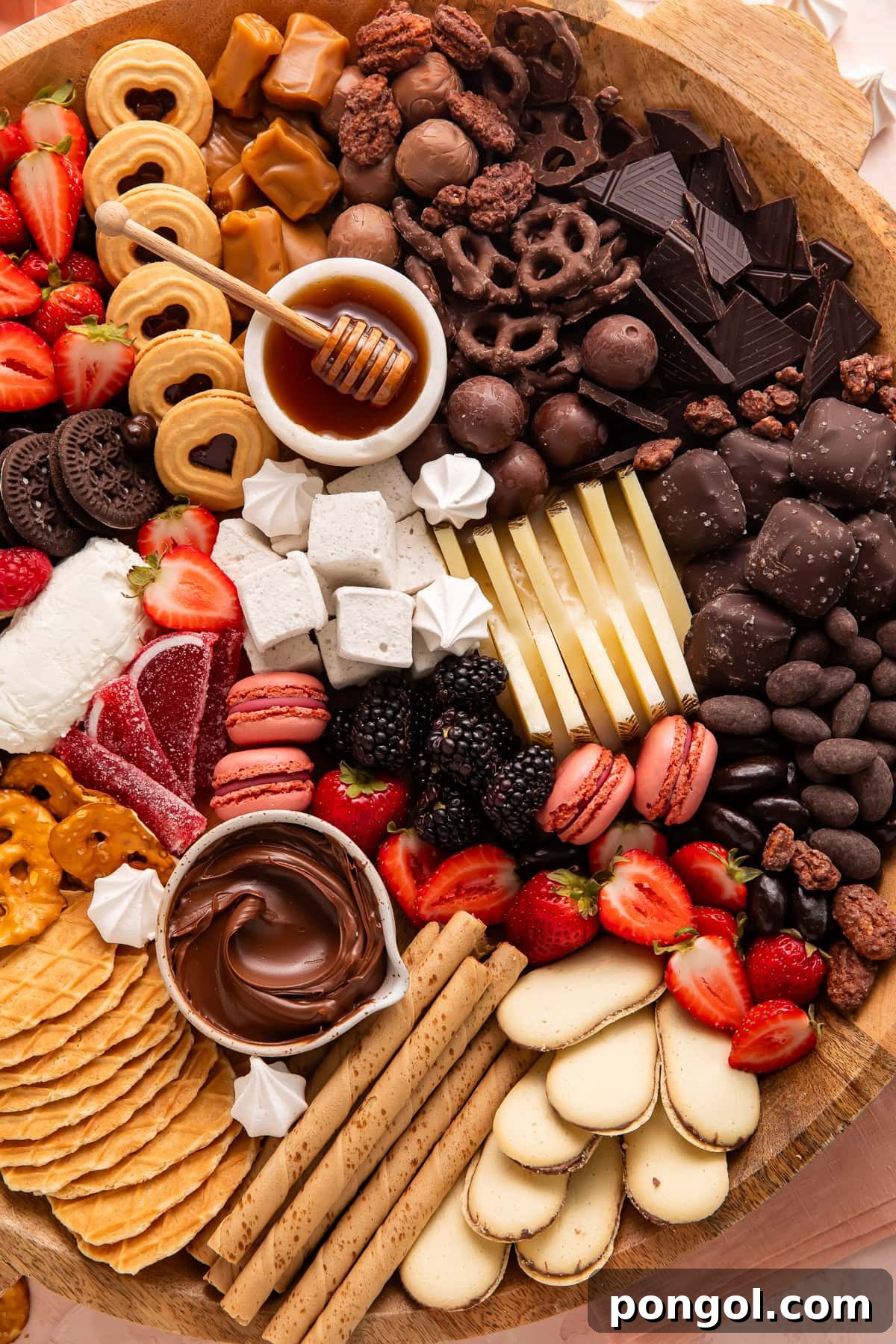 Section of dessert board featuring dark chocolate pieces, chocolate truffles, chocolate-covered pretzels, chocolate sandwich cookies, honey bowl, caramels, pirouette cookies, and fresh strawberries.