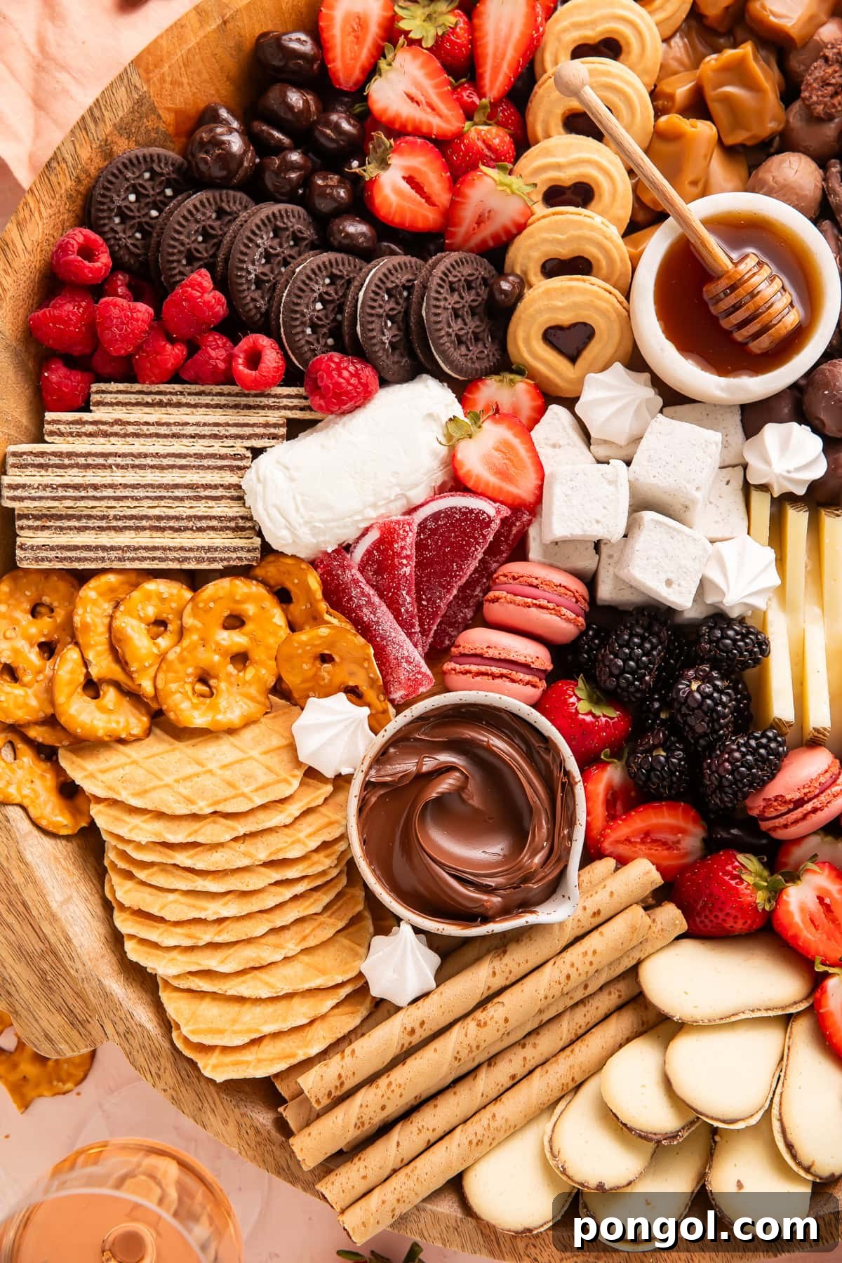 Close-up view of dessert board showing Oreos, waffle cookies, fresh strawberries, raspberries, honey bowl with dipper, chocolate dip, marshmallows, and pink macarons on wooden platter.