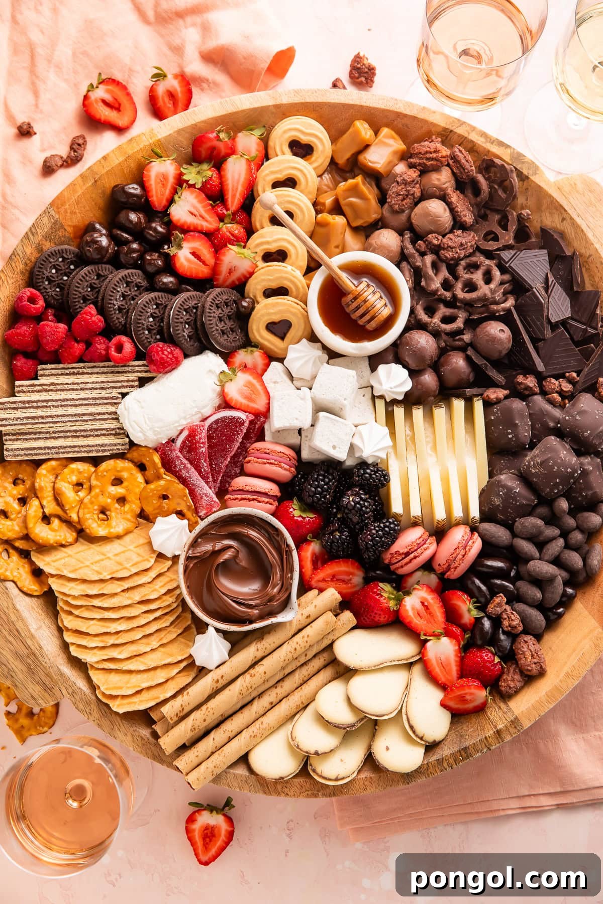 Full dessert charcuterie board on wooden platter with wine glasses and strawberries in background, featuring chocolates, cookies, berries, cheese, honey, and assorted sweets.