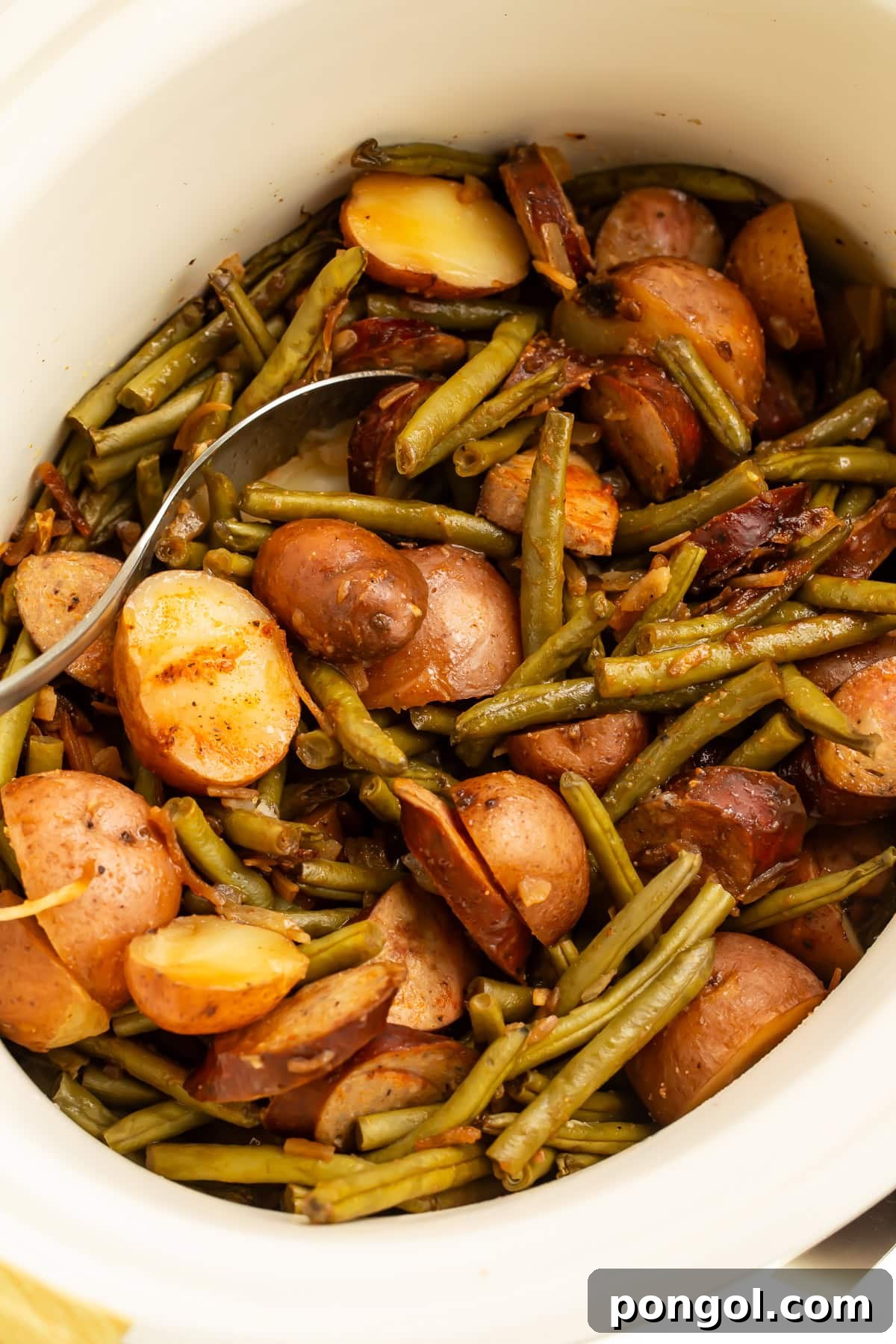 White slow cooker filled with cooked swamp potatoes showing tender red potatoes, green beans, and sliced sausage with serving spoon