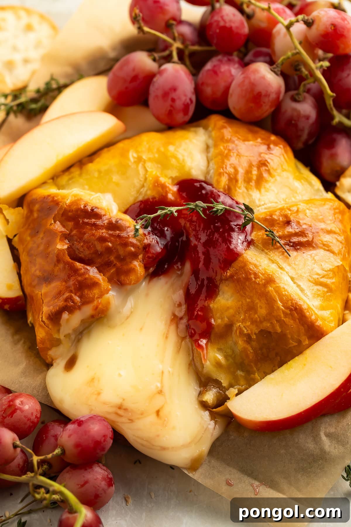 Golden brown brie en croute with melted cheese oozing out, topped with raspberry jam and fresh thyme, surrounded by grapes and apple slices.