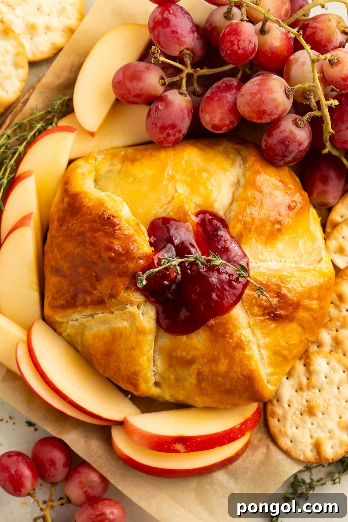 Whole baked brie en croute with golden puff pastry before cutting, topped with raspberry jam, served with crackers and fruit.