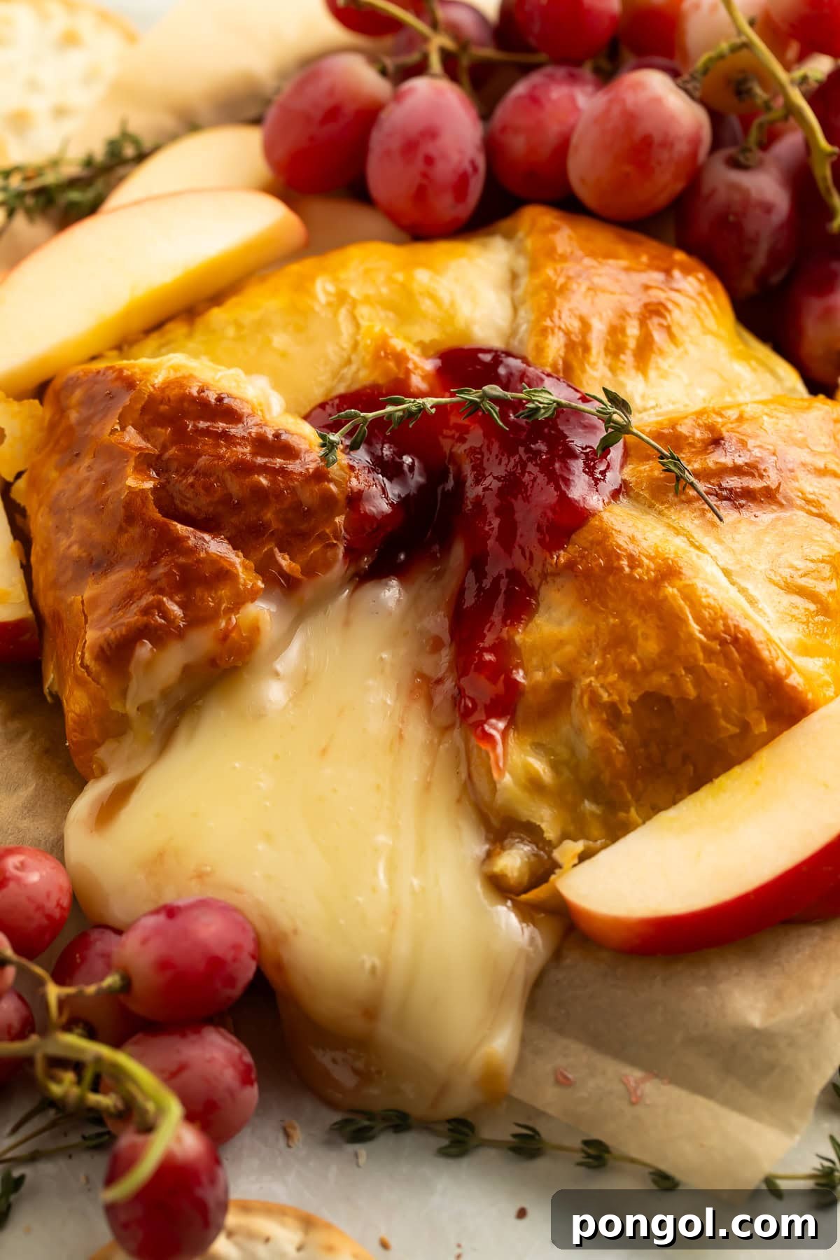 Close-up of sliced brie en croute showing melted brie cheese center with jam, flaky golden pastry exterior, grapes and apple slices.
