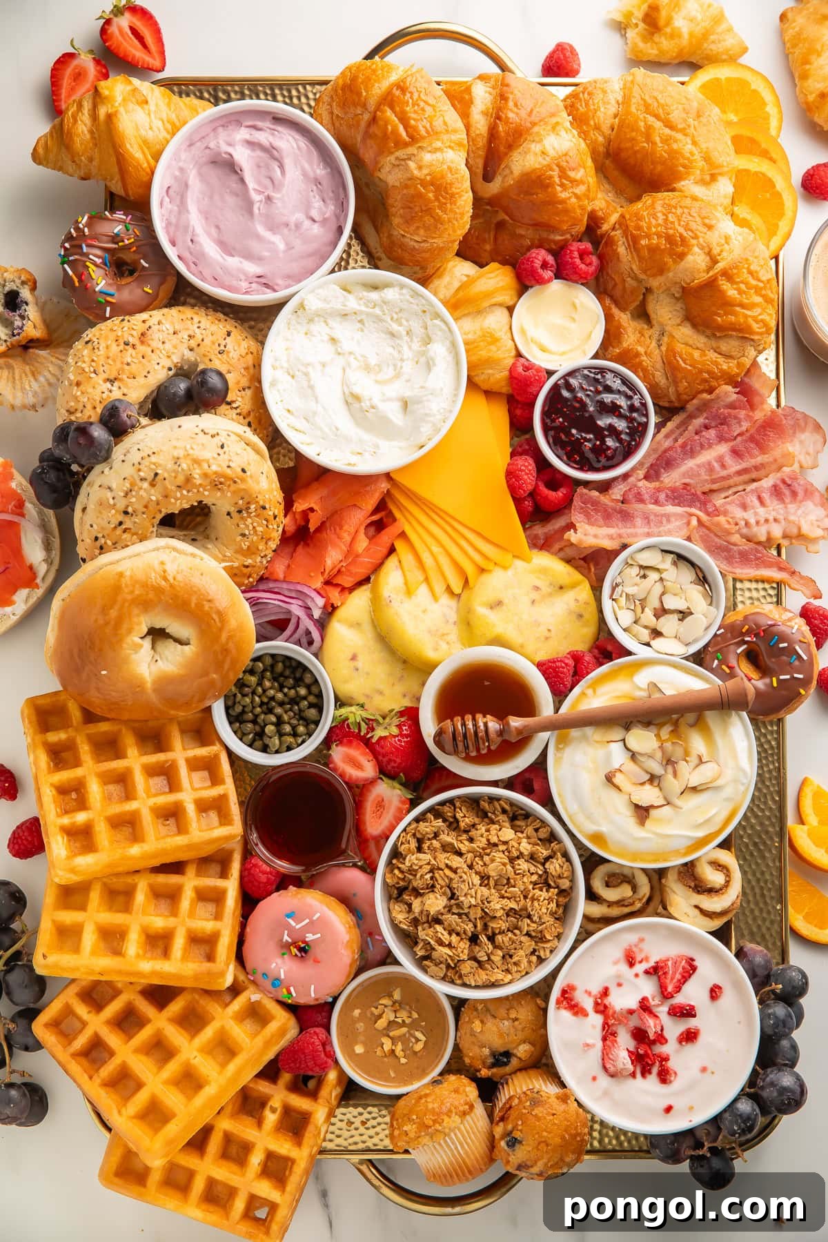 A beautifully arranged breakfast charcuterie board on a wooden tray, featuring an assortment of bagels, croissants, waffles, yogurt, crispy bacon, various cheeses, colorful fruits, fluffy muffins, and sweet donuts.