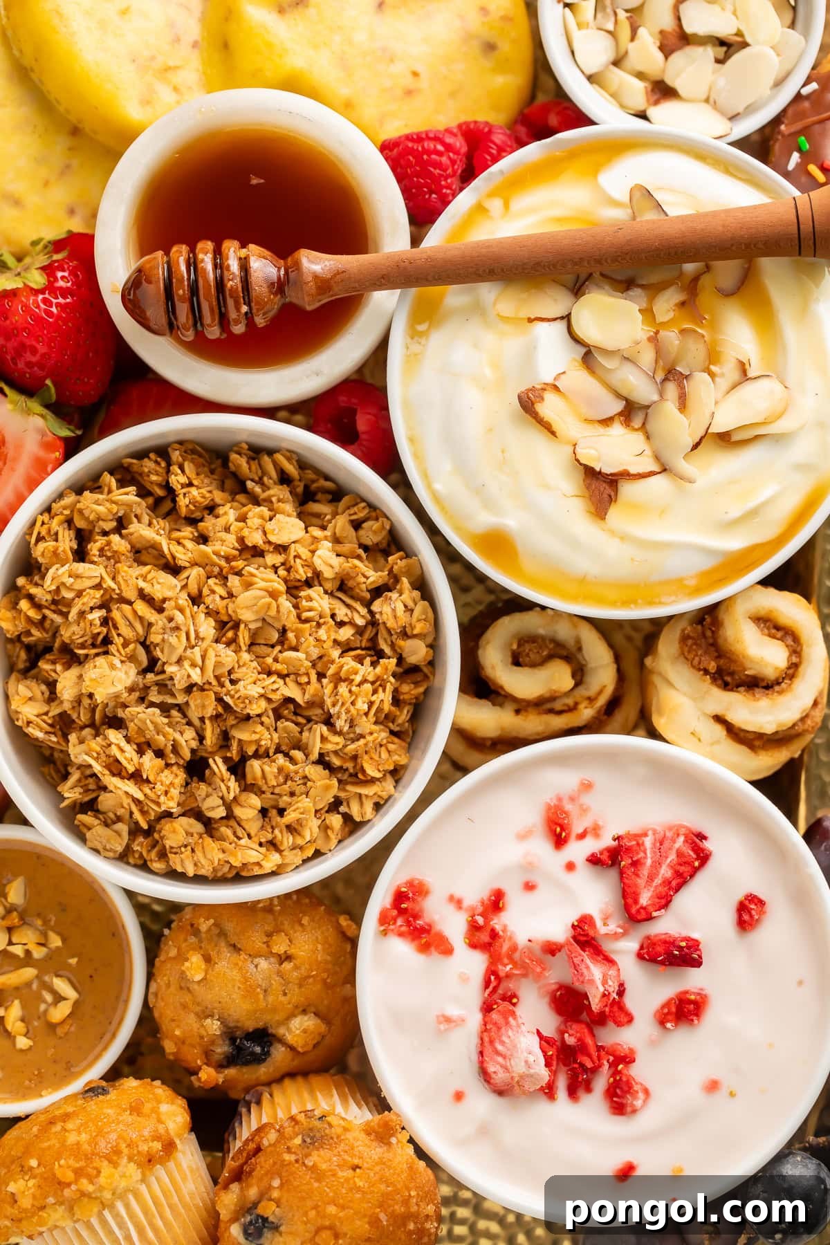 An overhead view of a vibrant breakfast charcuterie board, highlighting individual yogurt bowls, crunchy granola, an array of fresh berries, golden waffles, soft bagels, delightful muffins, and a drizzle of honey on a rustic wooden surface.