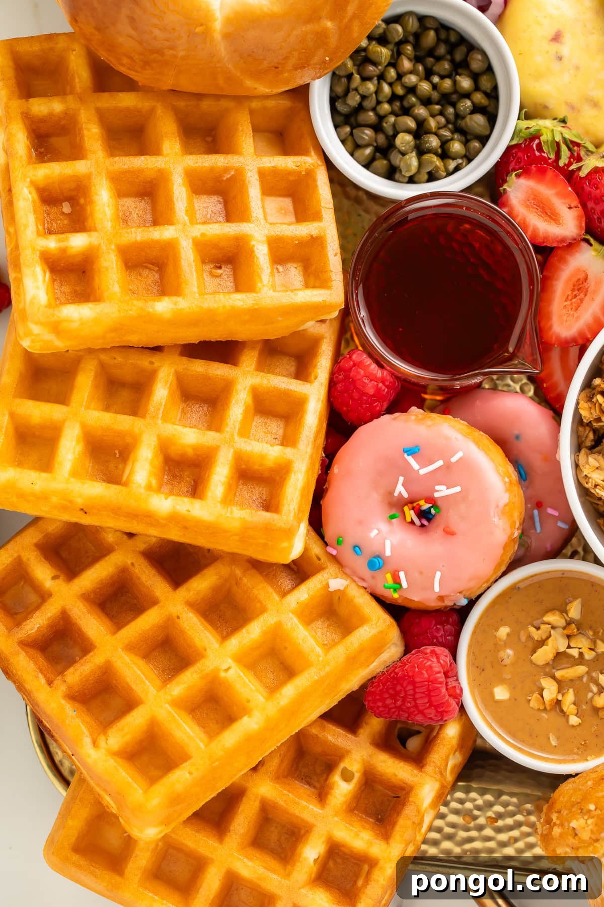 A close-up of a breakfast charcuterie board, highlighting perfectly golden Belgian waffles, a vibrant pink frosted donut, tiny briny capers, and juicy fresh strawberries.