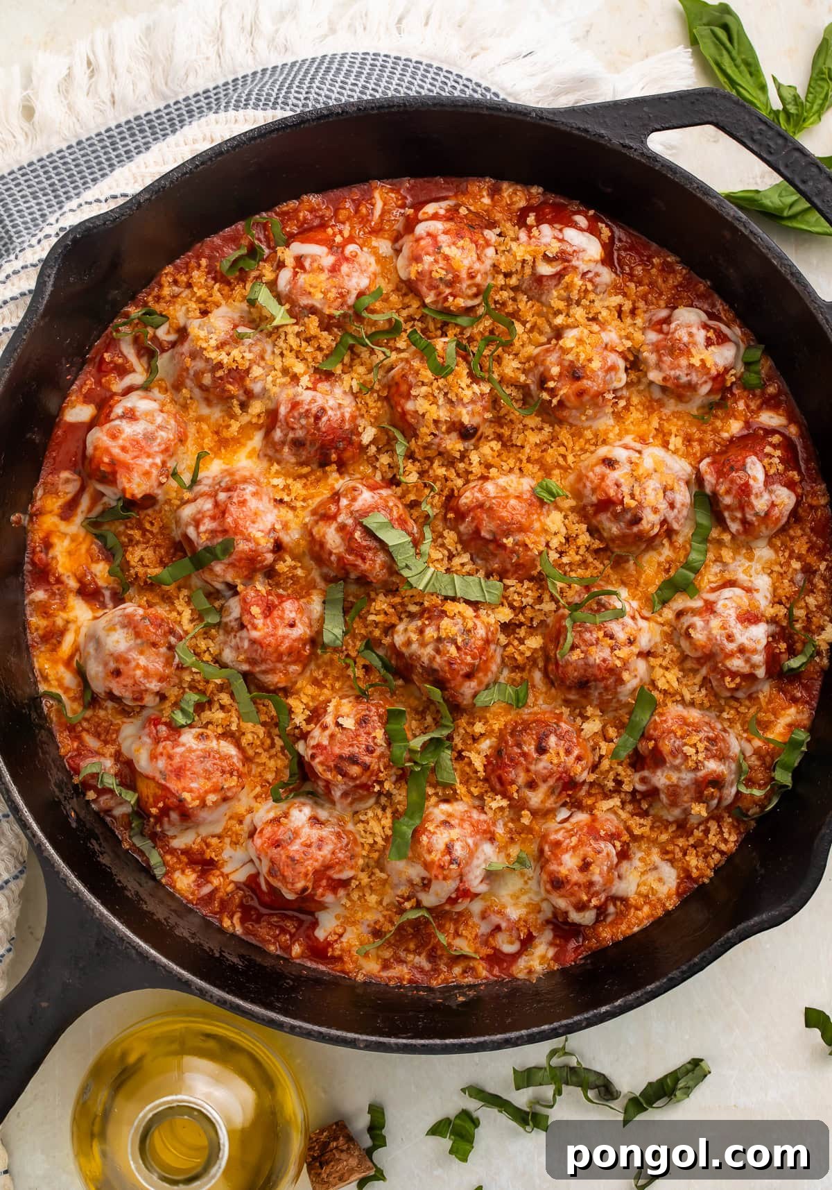 Full cast iron skillet of chicken parmesan meatballs with golden breadcrumbs, fresh basil, and olive oil on white countertop.