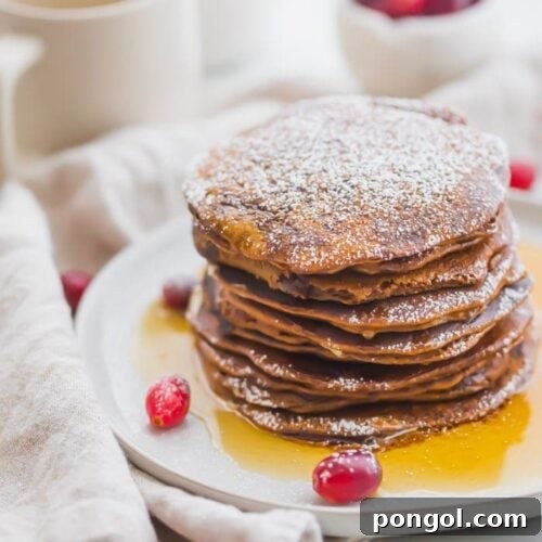 Stack of paleo gingerbread pancakes on a white plate with maple syrup around the bottom and fresh cranberries as garnish