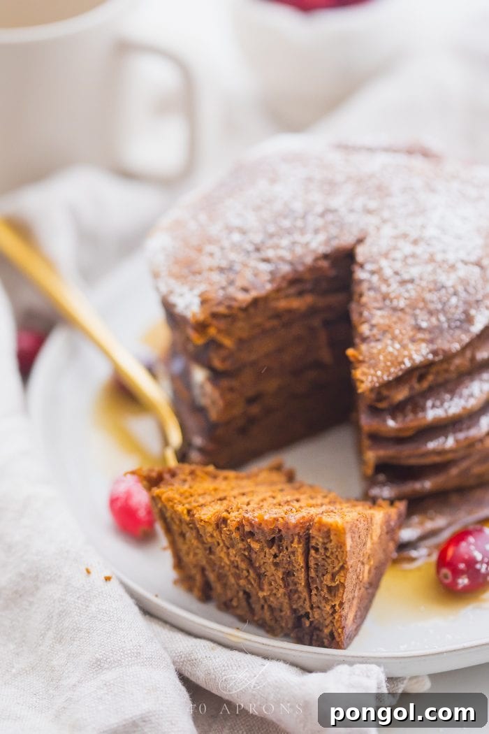 Stack of paleo gingerbread pancakes on a white plate with maple syrup around the bottom and a big bite cut out and fresh cranberries as garnish