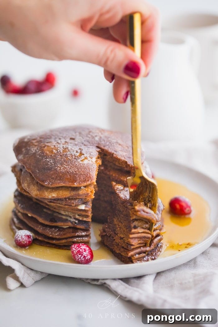 Stack of paleo gingerbread pancakes on a white plate with maple syrup around the bottom and a big bite cut out with a gold fork held between fingers in the piece and fresh cranberries as garnish