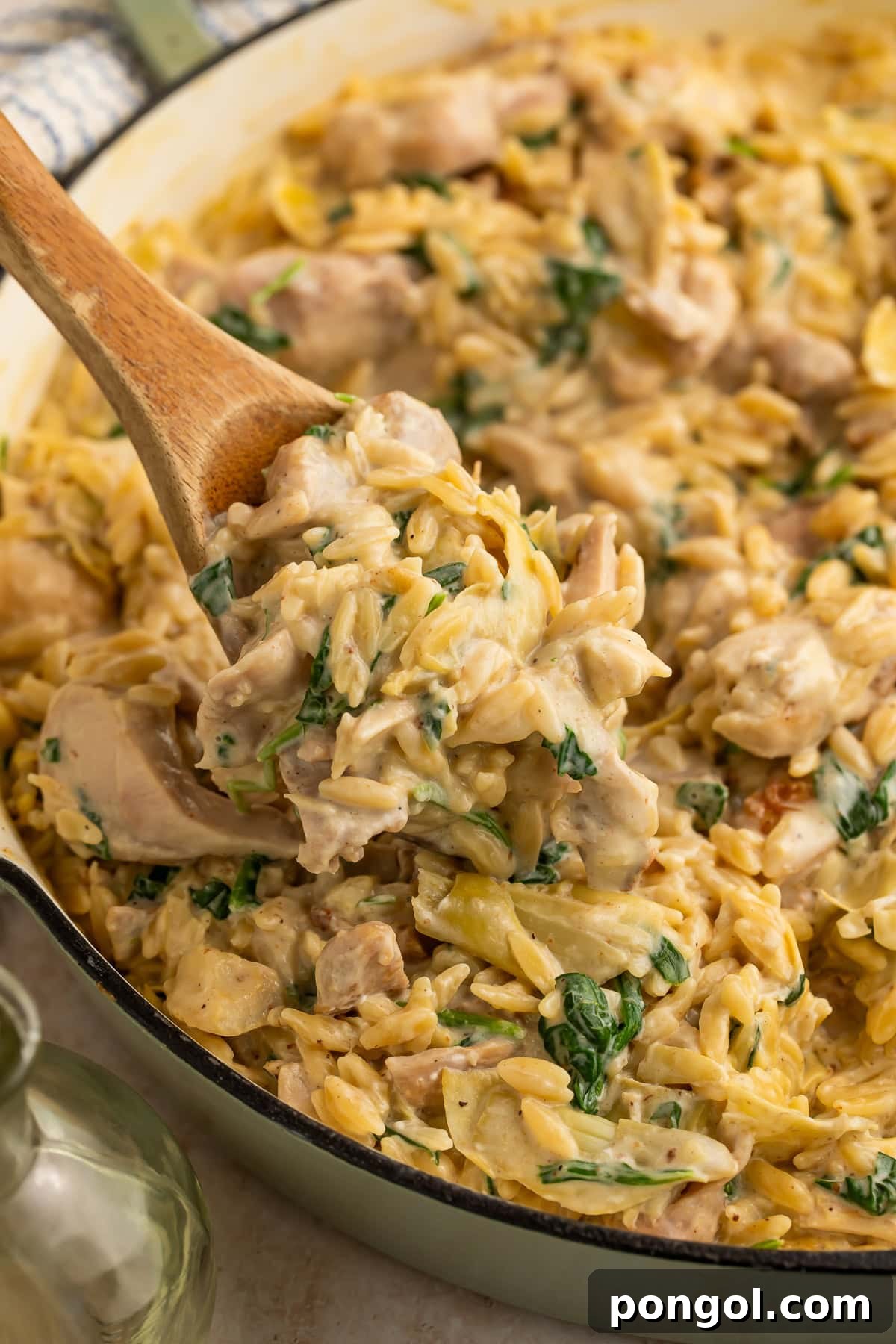 One Pan Creamy Spinach Artichoke Chicken Orzo 3 Wooden spoon lifting creamy chicken orzo pasta with spinach and artichokes from white skillet