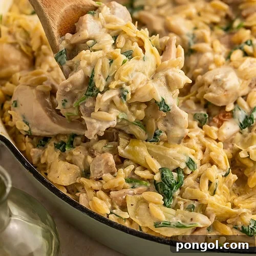 One Pan Creamy Spinach Artichoke Chicken Orzo 5 Wooden spoon lifting creamy chicken orzo pasta with spinach and artichokes from white skillet