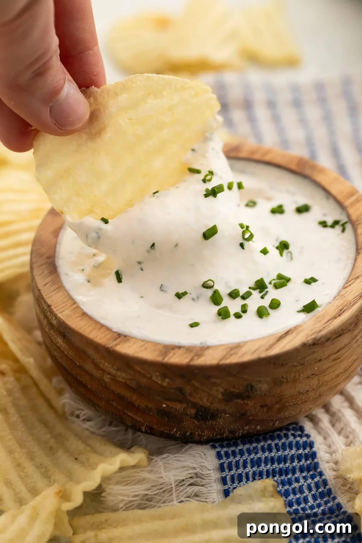 Hand holding ridged potato chip dipping into creamy herb dip in wooden bowl with fresh chives.
