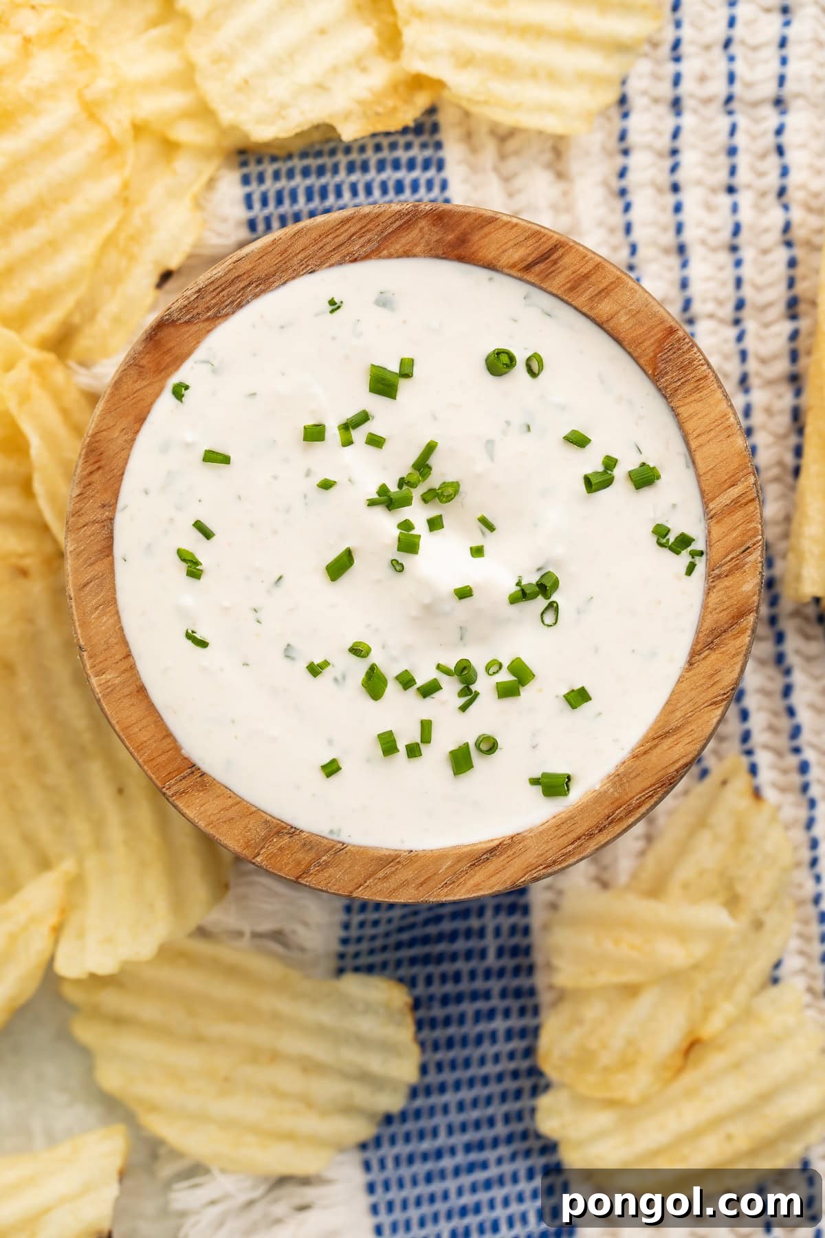Overhead view of creamy herb dip with fresh chives in wooden bowl surrounded by potato chips.