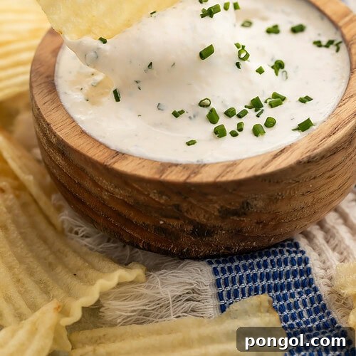 Hand holding ridged potato chip dipping into creamy herb dip in wooden bowl with fresh chives.