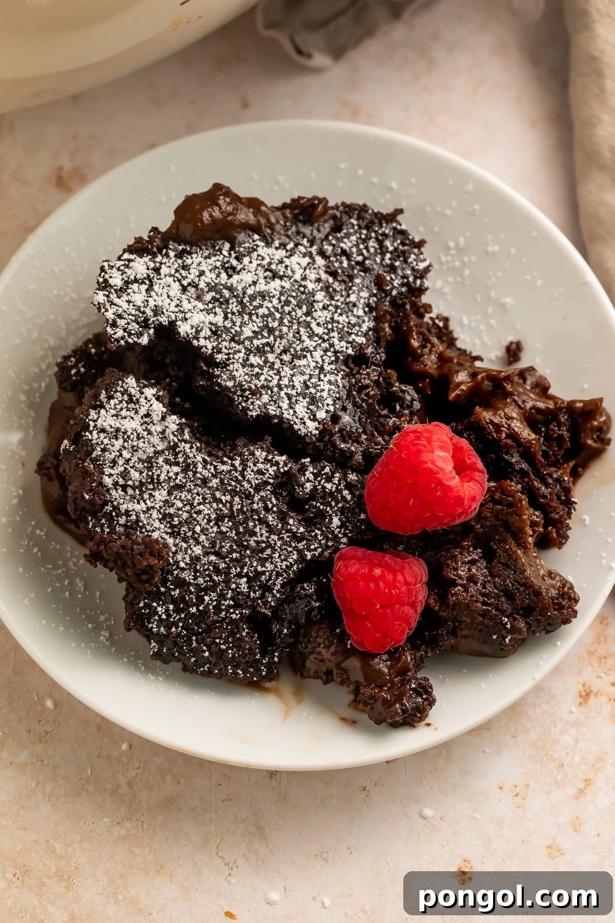 Close-up of a portion of chocolate lava cake with powdered sugar and raspberries on a white plate, highlighting the gooey center.