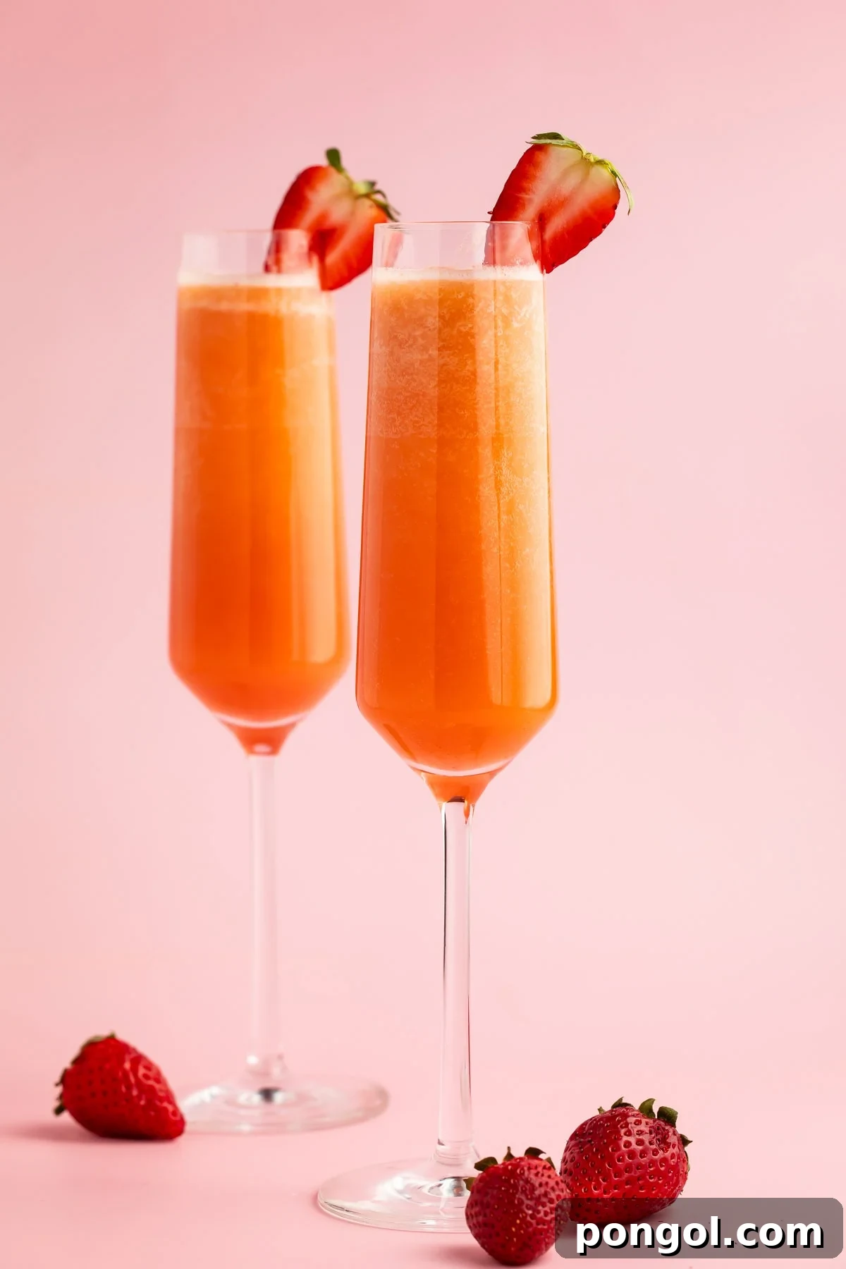 Two strawberry mimosas in champagne flutes garnished with halved strawberries on a pink background