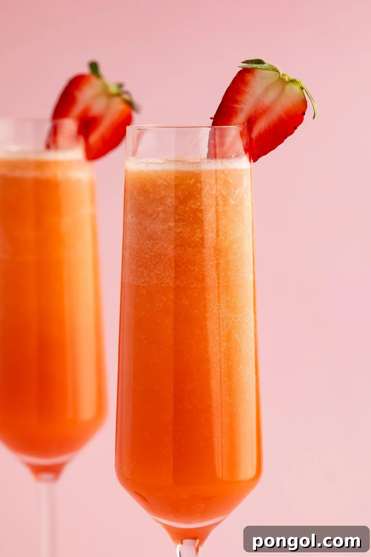 Close-up of two strawberry mimosas in champagne flutes garnished with fresh strawberry slices on pink background