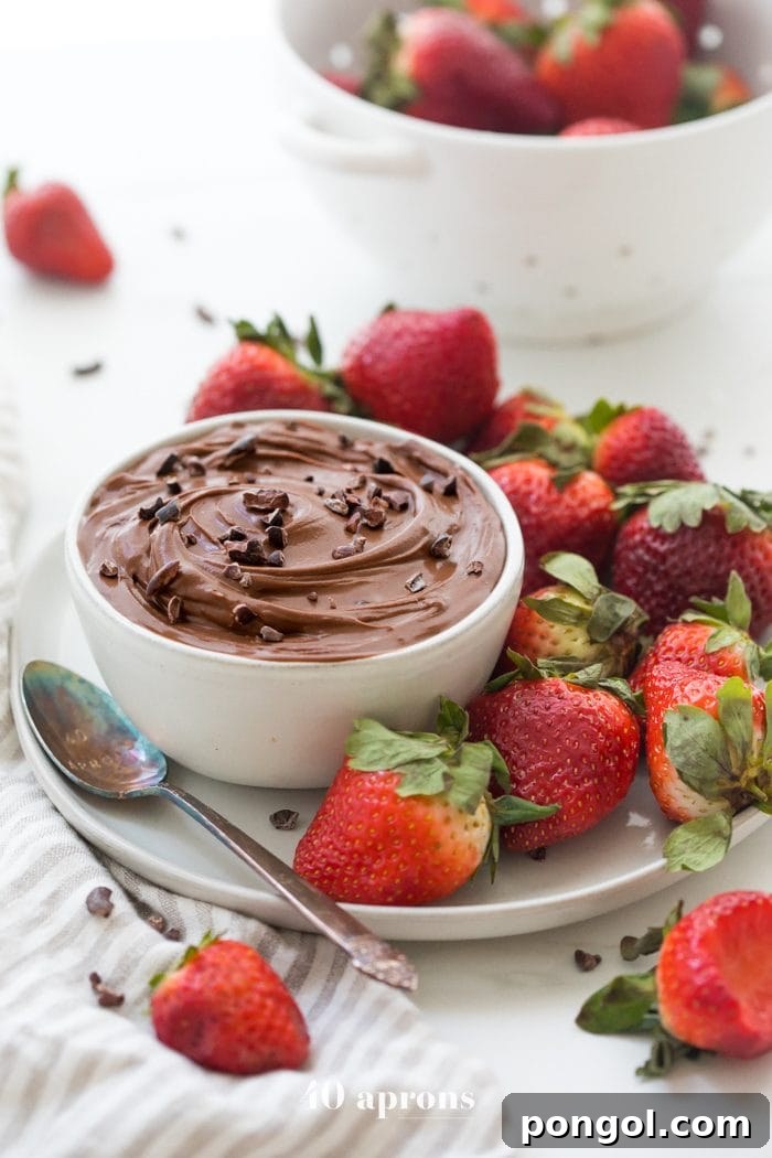 Close-up of a bowl of paleo chocolate mousse dip, showing its smooth and thick consistency, ready for dipping. A single strawberry is partially dipped, highlighting the delicious pairing.