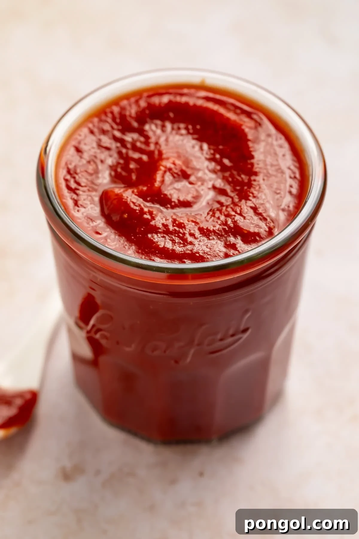 Glass jar of homemade ketchup with a white ketchup-dipped spoon off to one side.