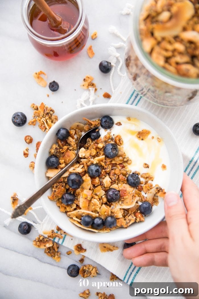 Hand holding a bowl of the crunchy paleo granola recipe over yogurt, topped with blueberries and a drizzle of honey